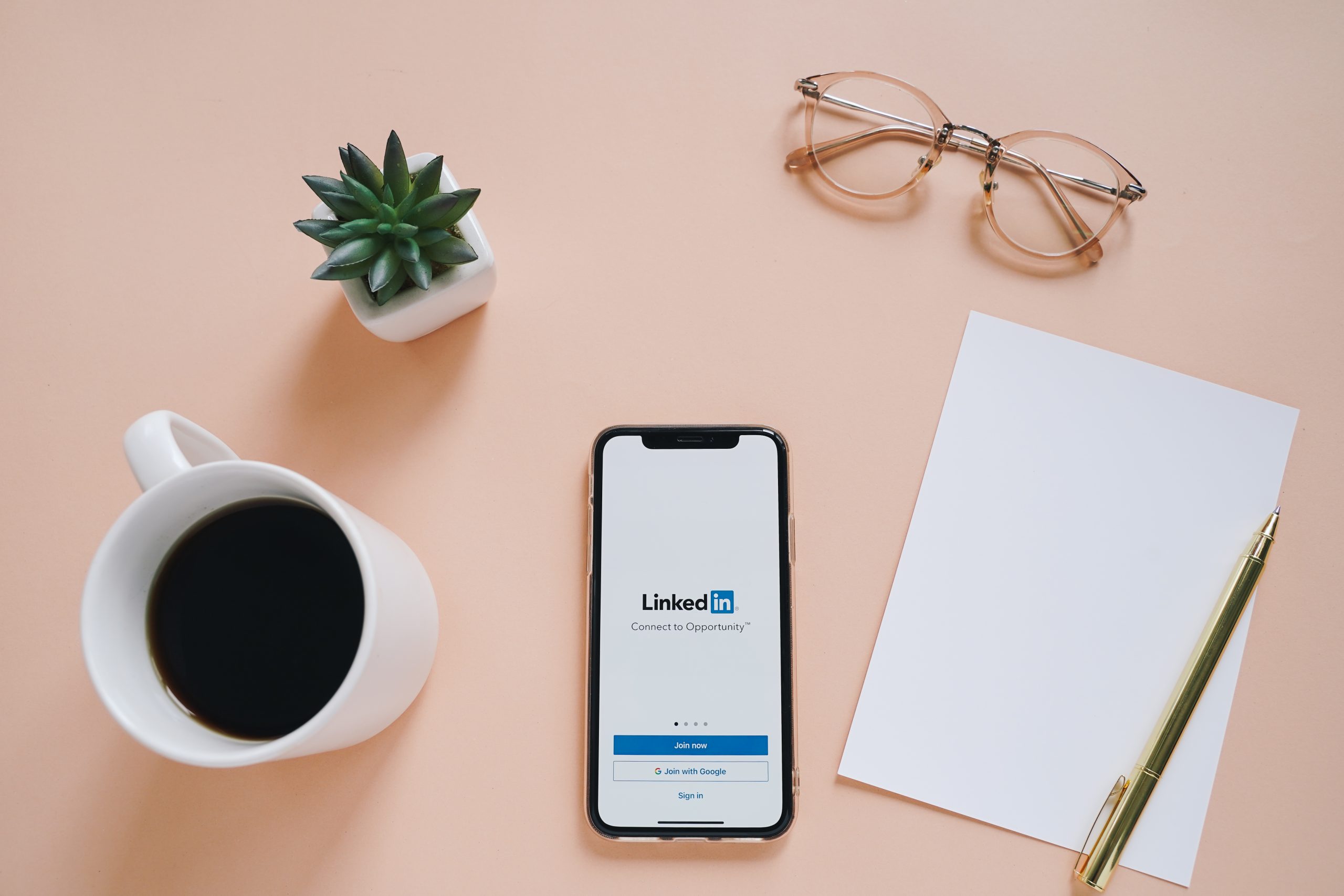 table with coffee, plant, glasses, paper, pen and cell phone turned on to LinkedIn app on it.