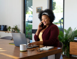 african american woman leader sitting at table, using laptop and talking on smartphone, working from home